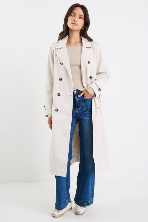 Woman wearing a beige trench coat, beige top, and blue jeans on a white background