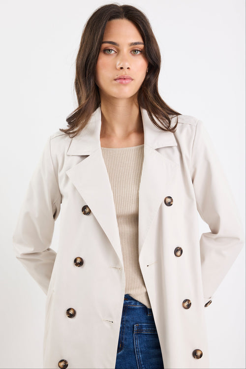 Woman wearing a beige trench coat over a beige sweater and blue jeans on a white background
