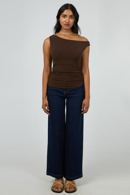 Woman wears a fitted, sleeveless chocolate brown top with an off-shoulder neckline, paired with dark denim jeans and minimal jewellery for a sleek, modern vibe.