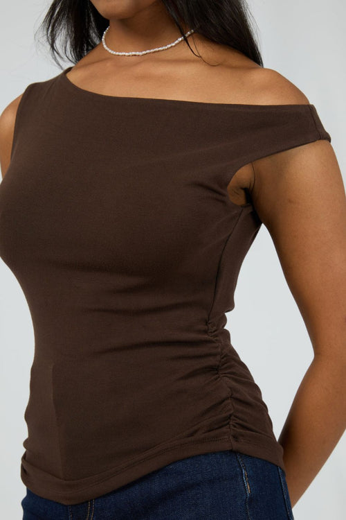 Woman wears a fitted, sleeveless chocolate brown top with an off-shoulder neckline, paired with dark denim jeans and minimal jewellery for a sleek, modern vibe.