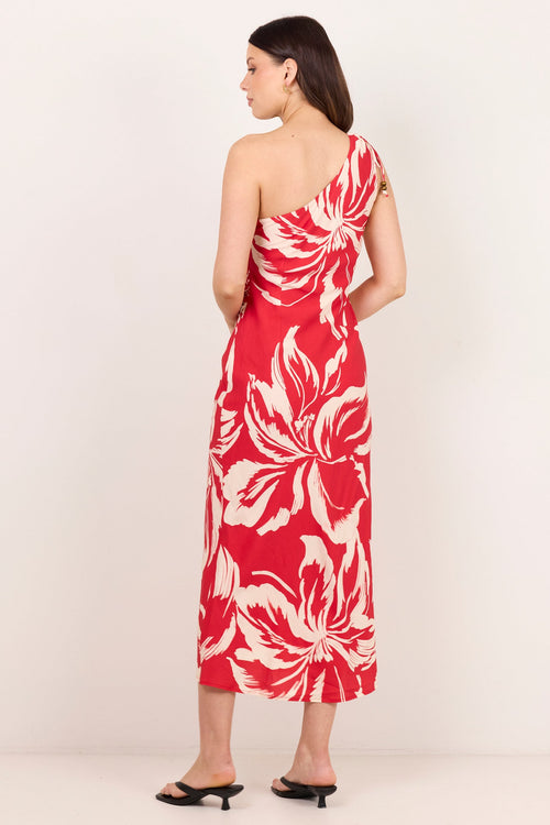 A woman modeling a one-shoulder midi dress with a bold red and cream floral print, shown from a side-back angle against a plain off-white studio background.