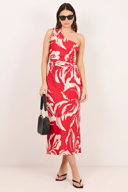 A woman modeling a red one-shoulder midi dress with a bold white floral pattern, styled with black sunglasses, a black handbag, and black sandals against a plain off-white studio background.