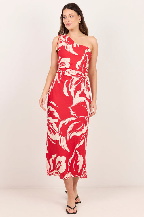 A woman modeling a one-shoulder midi dress with a bold red and cream floral pattern, standing against a plain off-white studio background.