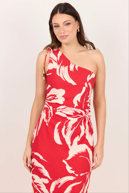 A woman modeling a red one-shoulder dress with a bold white floral print against a plain light beige studio background.