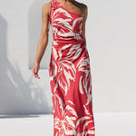 A woman models a red and white tropical leaf print maxi dress in an outdoor setting next to a swimming pool, with a white wall and green trees in the background.