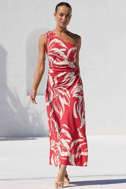 A woman models a red and white tropical leaf print maxi dress in an outdoor setting next to a swimming pool, with a white wall and green trees in the background.
