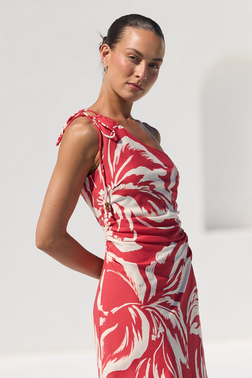 A woman modeling a red one-shoulder maxi dress with a white tropical floral print, set against a bright white minimalist background.