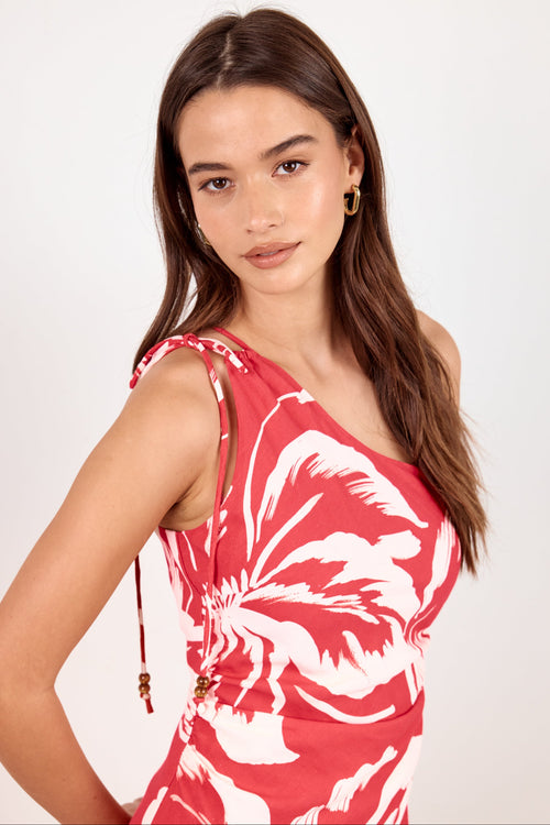A woman modeling a red and white tropical floral print one-shoulder dress against a plain white studio background.