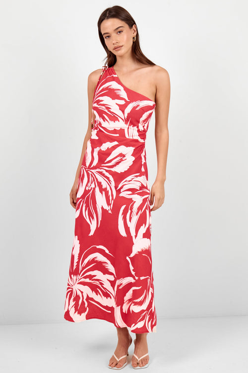 A woman modeling a red one-shoulder maxi dress with a white tropical floral print, standing against a light gray studio background.