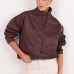 A woman modeling a cropped, high-neck jacket in a dark brown satin-like fabric, paired with cream-colored trousers, against a plain white studio background.