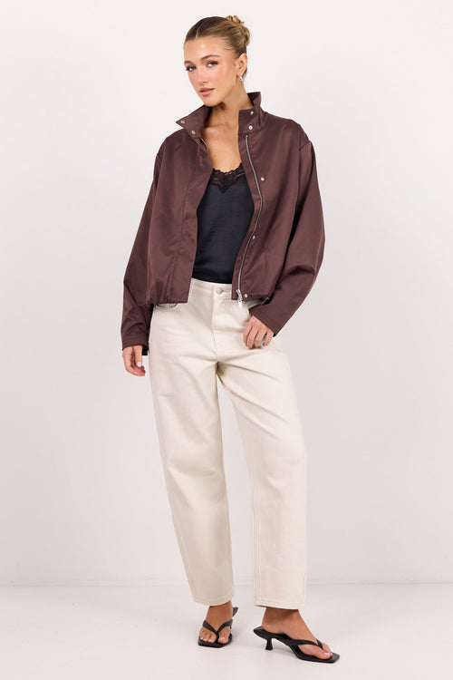 A woman models a dark brown satin bomber jacket over a black top and cream-colored wide-leg trousers, standing against a plain white studio background.