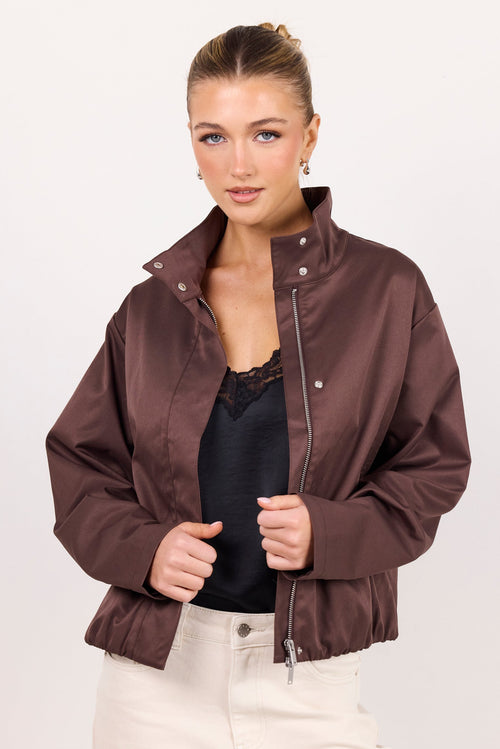 A woman modeling a brown satin bomber jacket over a black top and cream pants, photographed against a plain light gray studio background.