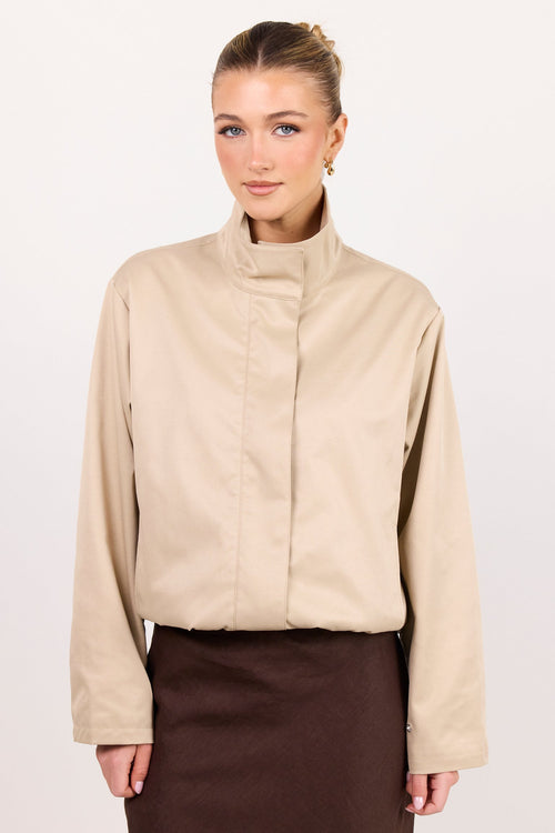 A woman modeling a beige long-sleeve mock-neck jacket with a unique front seam detail, paired with a dark brown skirt, against a plain white studio background.