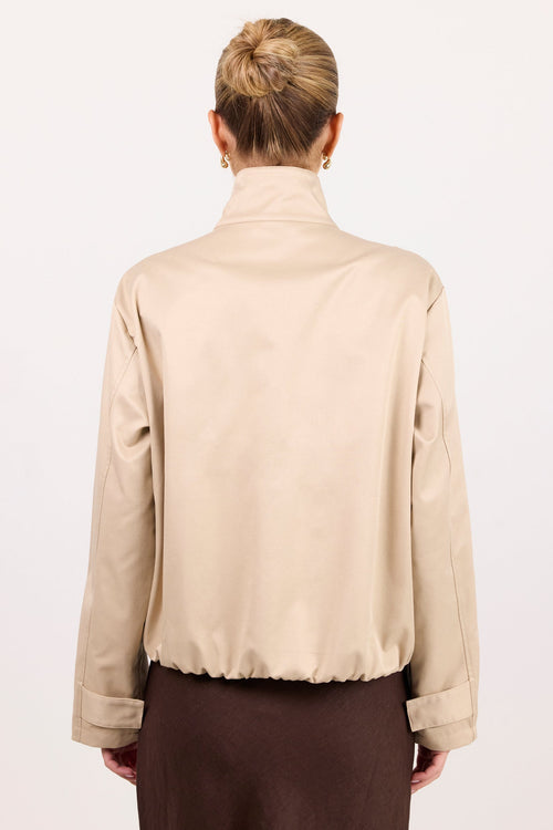A woman viewed from behind wearing a beige long-sleeve mock-neck jacket and a dark brown skirt, set against a plain white studio background.
