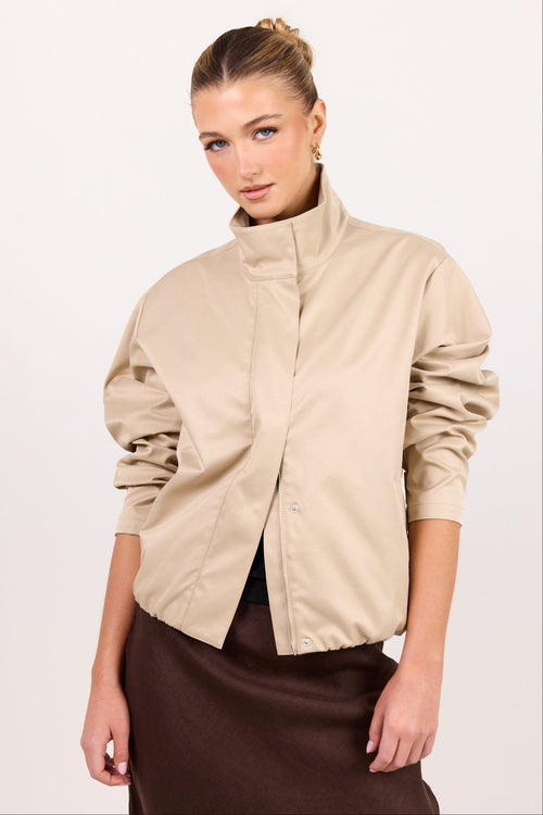 A woman modeling a beige wrap-style jacket with a high stand collar and gathered sleeves, paired with a dark brown skirt, against a plain white studio background.