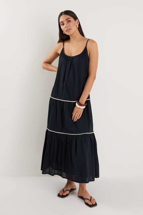 Woman wearing a plain black tiered maxi dress with strappy sleeves, white trim details, and back tie feature, styled with black sandals.