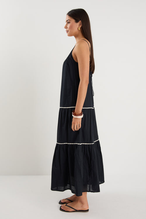 Woman wearing a plain black tiered maxi dress with strappy sleeves, white trim details, and back tie feature, styled with black sandals.