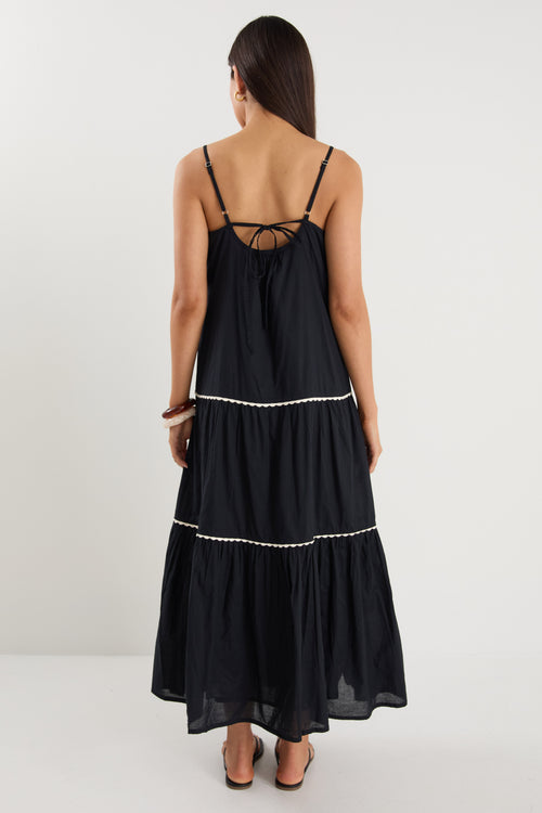 Woman wearing a plain black tiered maxi dress with strappy sleeves, white trim details, and back tie feature, styled with black sandals.