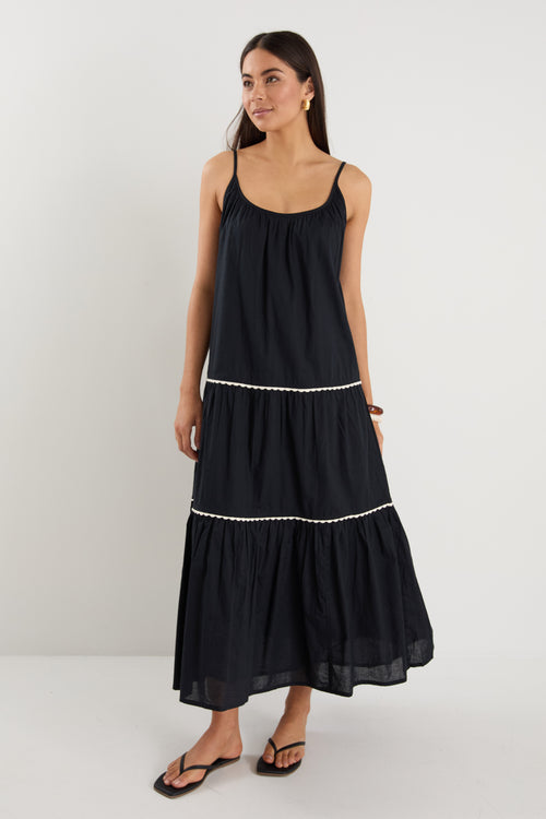 Woman wearing a plain black tiered maxi dress with strappy sleeves, white trim details, and back tie feature, styled with black sandals.