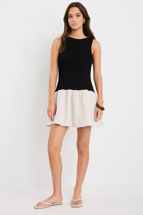 Woman wearing a sleeveless mini dress with a black shirred bodice and a natural beige flared skirt, styled simply for a casual chic look.