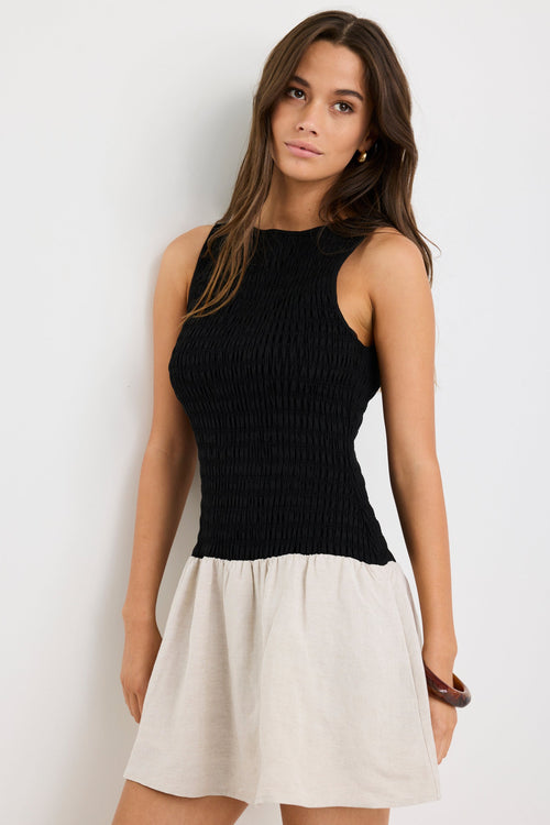 Woman wearing a sleeveless mini dress with a black shirred bodice and a natural beige flared skirt, styled simply for a casual chic look.