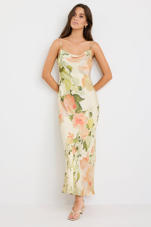 Woman wearing a cream bias-cut slip dress with peach floral print, featuring thin straps, a cowl neckline, and tie-back detail, styled with minimal accessories for a romantic look.