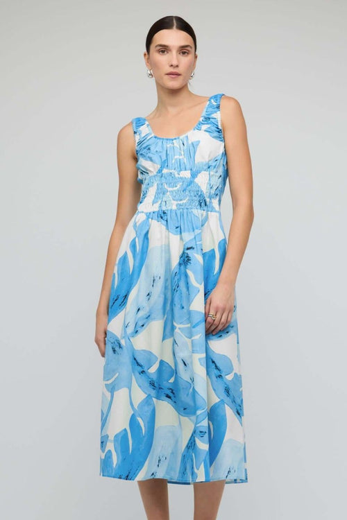 Woman wearing a light blue and white sleeveless midi dress with a shirred waist and soft leaf print, featuring a relaxed, floaty silhouette.