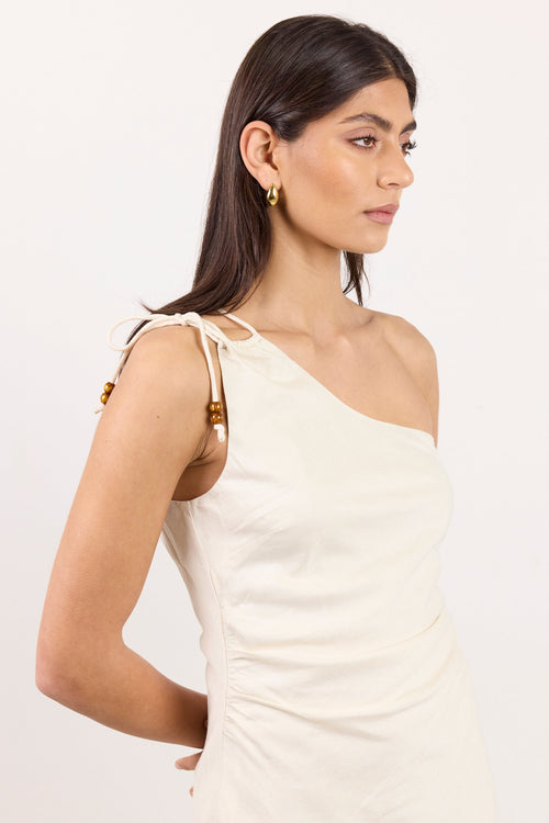 A woman modeling a cream-colored, one-shoulder wrap dress with gold-toned decorative accents on the shoulder straps, set against a plain white studio background.