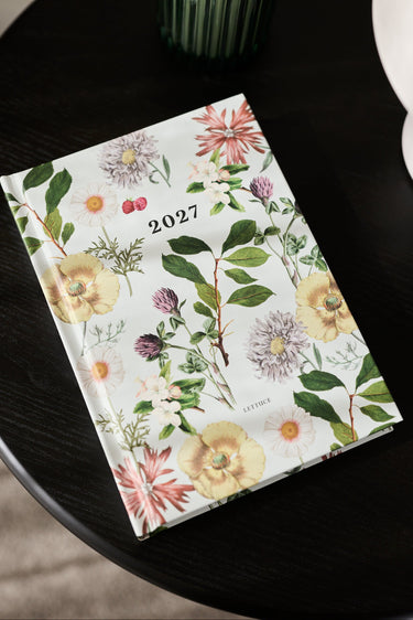 A 2027 floral planner or desk calendar lies open on a dark round table, showing a botanical cover with yellow flowers, green leaves, and small pink and purple blossoms. A green ribbed candle holder and part of a white object are visible in the background.
