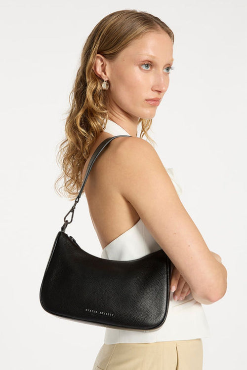 A sleek black leather crossbody bag with a soft, curved silhouette and pebble-grain texture. It features a detachable shoulder strap with silver hardware clasps and 