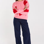 Woman wearing a pink sweater with red heart patterns and dark blue pants on a white background