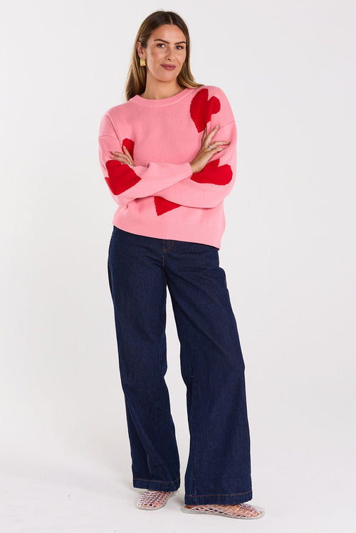 Woman wearing a pink sweater with red heart patterns and dark blue pants on a white background