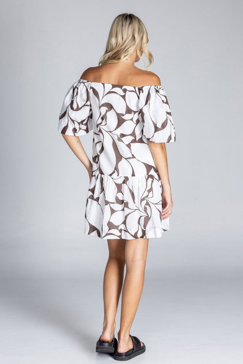 Woman wearing an off-the-shoulder white mini dress with abstract brown swirl print, puff sleeves, and a tiered skirt, standing against a plain grey background.