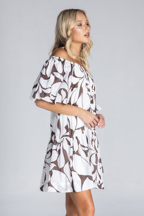 Woman wearing an off-the-shoulder white mini dress with abstract brown swirl print, puff sleeves, and a tiered skirt, standing against a plain grey background.