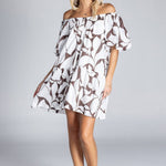 Woman wearing an off-the-shoulder white mini dress with abstract brown swirl print, puff sleeves, and a tiered skirt, standing against a plain grey background.