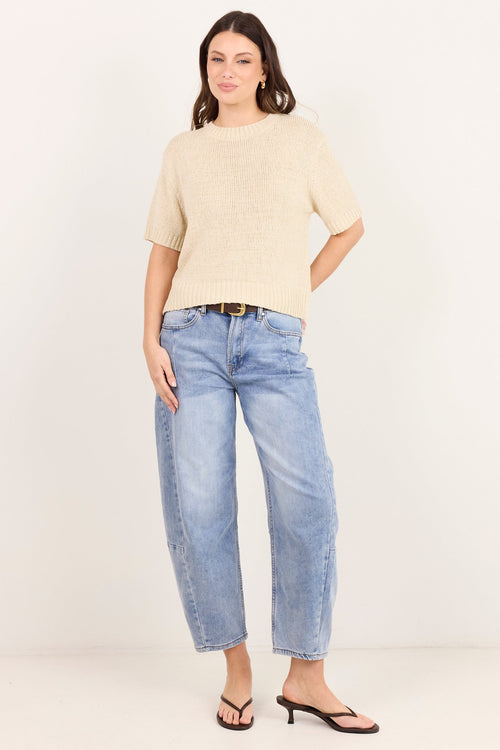 A woman modeling a beige short-sleeve knit top and light blue cropped jeans with black sandals, standing against a plain off-white studio background.