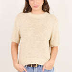A woman modeling a short-sleeve cream-colored ribbed knit sweater with light-wash blue jeans against a plain off-white background.