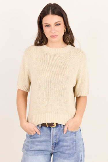 A woman modeling a short-sleeve cream-colored ribbed knit sweater with light-wash blue jeans against a plain off-white background.