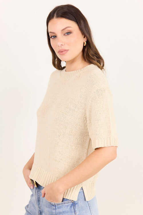 A woman wearing a short-sleeve cream-colored ribbed knit sweater and light blue jeans, standing against a plain off-white studio background.