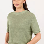 A woman modeling a short-sleeve knit sweater in sage green, paired with white pants and a brown belt, against a plain white studio background.