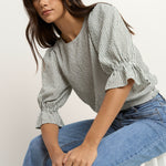 Woman wears a khaki and white striped puff sleeve top with frill detail, paired with light denim jeans.