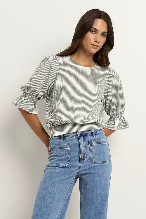 Woman wears a khaki and white striped puff sleeve top with frill detail, paired with light denim jeans.