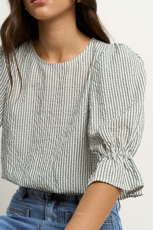 Woman wears a khaki and white striped puff sleeve top with frill detail, paired with light denim jeans.