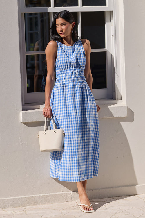 A woman stands outdoors in front of a white wall and window, wearing a sleeveless blue-and-white gingham midi dress with a gathered waist. She is holding a small white handbag and wearing white sandals.