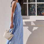 A woman wearing a blue and white gingham sleeveless maxi dress walks outdoors next to a white wall with a window, holding a light beige handbag.