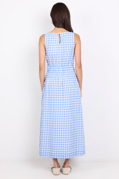 A woman viewed from behind wearing a sleeveless blue and white gingham maxi dress with a small keyhole cutout at the back, paired with beige flat shoes, against a plain white studio background.