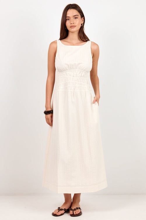 A woman modeling a sleeveless, off-white midi dress with a smocked bodice and side pockets, standing against a plain white studio background.