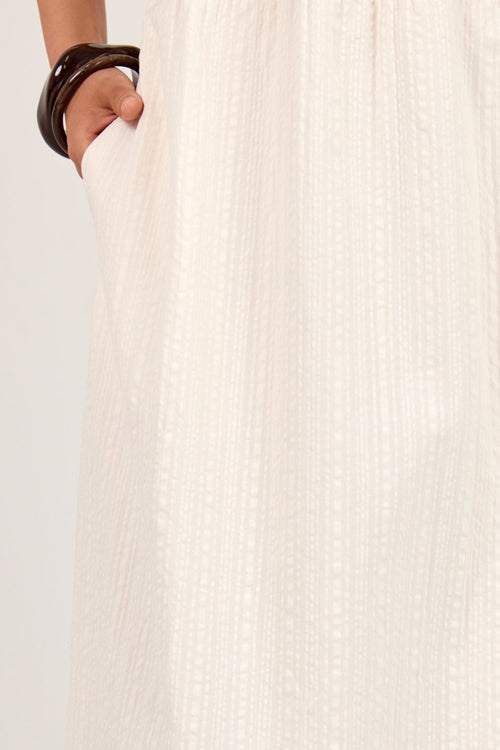 An off-white cotton gauze midi skirt with vertical textured stripes and side pockets, shown on a model wearing dark brown beaded sandals against a white studio background.
