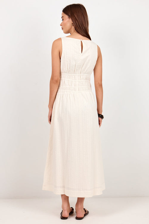 A woman viewed from the back wearing a sleeveless, off-white midi dress with a smocked waist and a small keyhole cutout at the back, paired with black flat sandals against a plain white studio background.