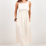A woman modeling a sleeveless, off-white midi dress with a smocked bodice and side pockets, paired with black sandals, against a plain white studio background.